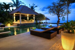 A poolside area with lounge chairs, a thatched-roof structure, and a view of the ocean. - 5 BR Luxurious Beachfront Villa - Large Swimming Pool &  Staff - Lovina Beach (Dencarik)