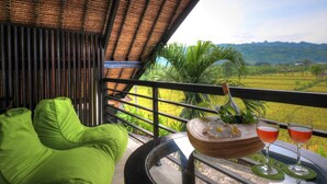 Un balcón con una silla acolchada verde, una mesa trenzada con una botella y vasos, y vistas a un paisaje verde.