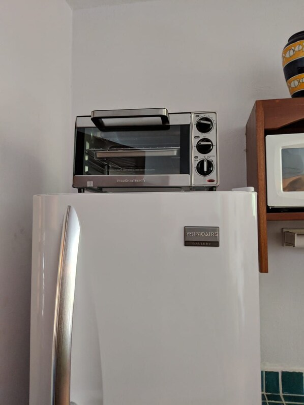 A kitchen with a built-in toaster oven on top of a white refrigerator, a microwave to the right, and a wooden cabinet above the microwave.