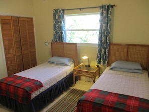 A room with two single beds, a wooden wardrobe, a small bedside table with a lamp, and a window with patterned curtains.