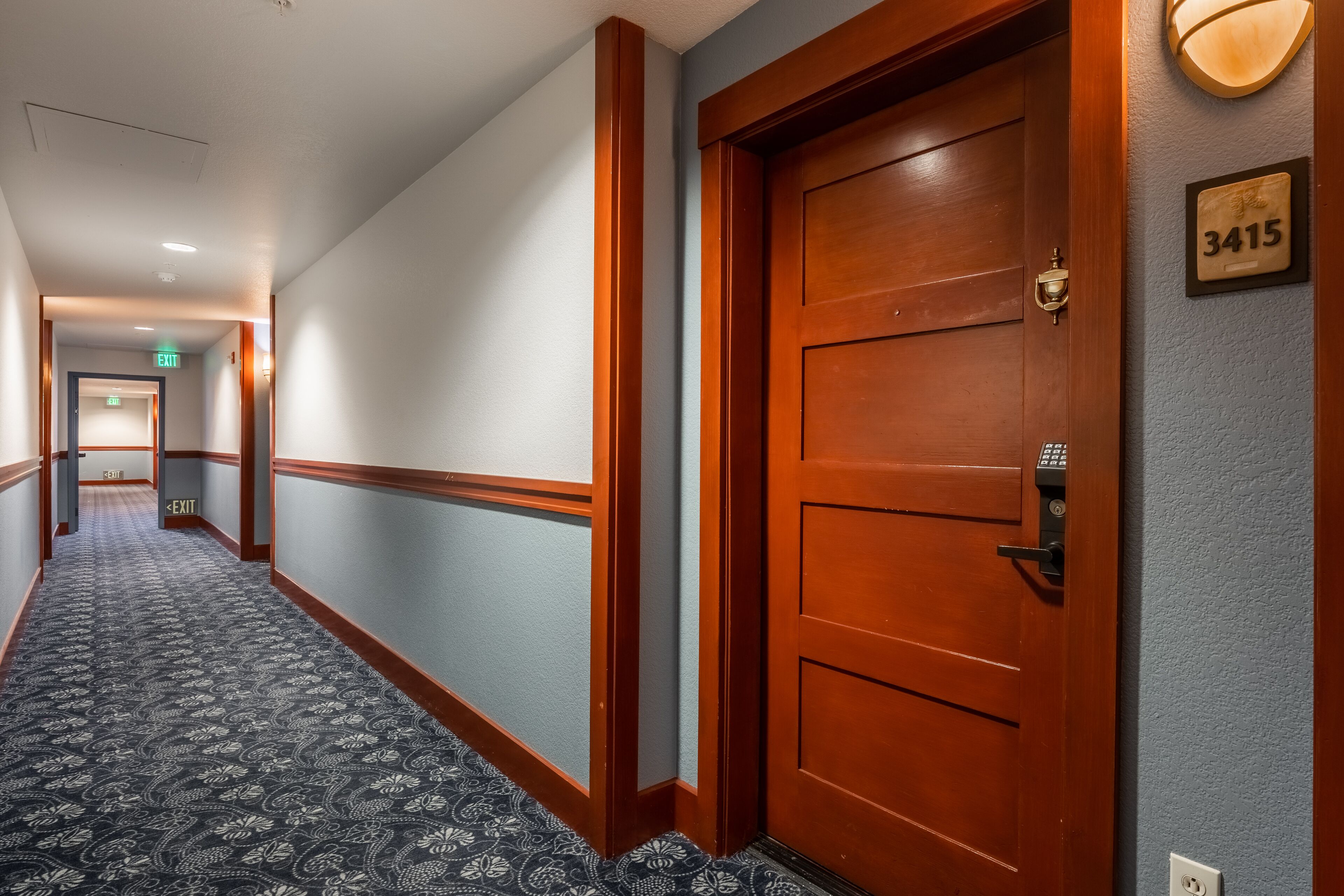 A hotel hallway with a patterned carpet, a wooden door with a number plate, and an exit sign.