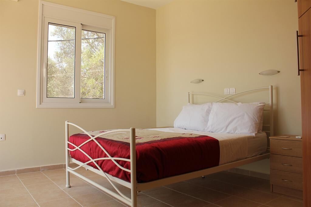 A single bed with a metal frame, a red and white bedspread, and white pillows. There is a window with a view of trees, a bedside table, and a wooden cabinet.