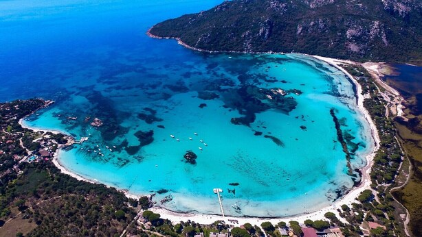 Ein Luftbild einer Küstenlandschaft mit klarem türkisblauem Wasser, einem Sandstrand und einer kleinen Insel mit dichter Vegetation.