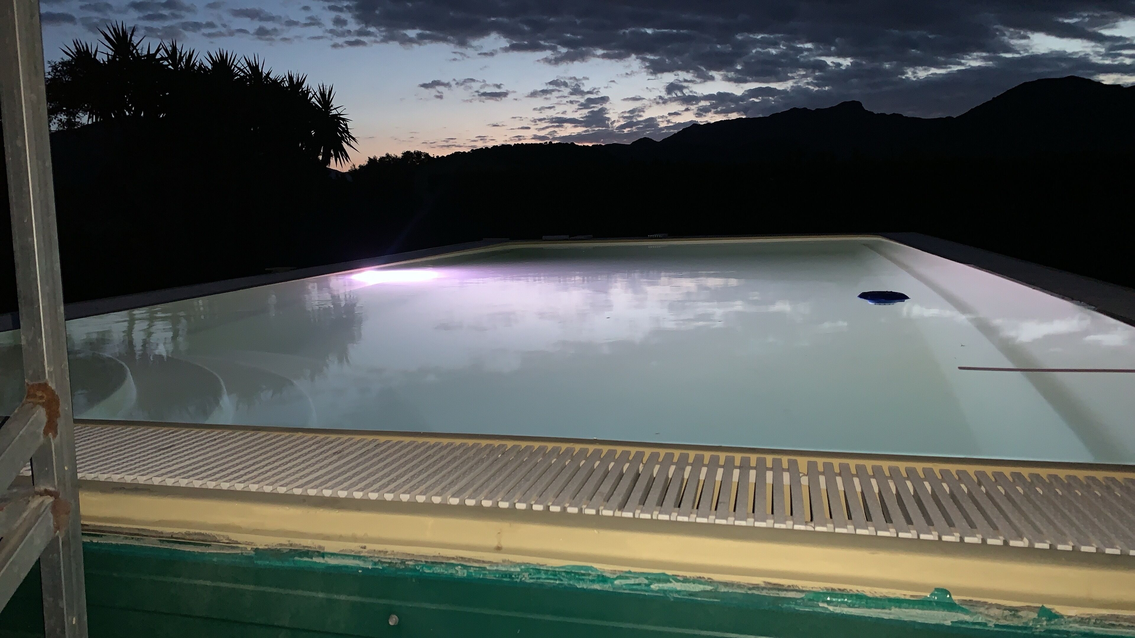 A swimming pool with clear water, surrounded by a metal deck and a view of mountains and trees at dusk.