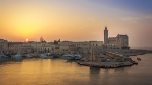 Une ville côtière dotée de bâtiments historiques, d’une tour imposante et de bateaux amarrés dans le port au coucher du soleil.