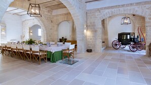 Une salle à manger avec une longue table dressée pour un repas, entourée de chaises en bois. On aperçoit une voiture à cheval garée au fond.