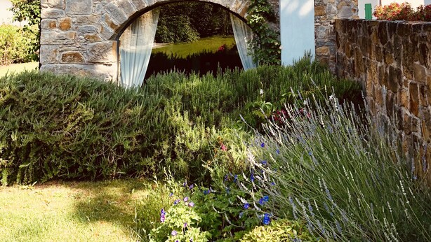 Un edificio in pietra con persiana in legno, un camino e un giardino con diverse piante e fiori.