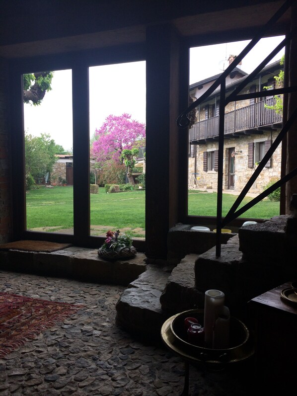 A room with a stone floor, a view of a garden with a pink flowering tree, and a balcony with wooden railings. - Casa Ponca, a splendid nest in the heart of the Friuli Collio (Cormons)