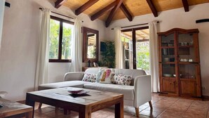 Ein Zimmer mit Holzboden, einem Sofa mit gemusterten Kissen, einem Holzschrank und einem Fenster mit Vorhängen.