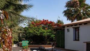 Un espace jardin agrémenté d’un panneau indiquant « PERLA DEL SUR », entouré de plantes tropicales et bordé d’un bâtiment blanc doté d’une fenêtre et d’un lampadaire.