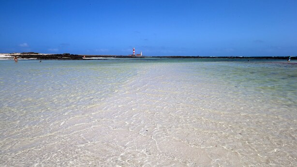 Une plage aux eaux claires et peu profondes, un phare à l’horizon et un paysage rocheux.