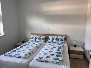 Una cama doble con edredón blanco y azul de lunares, dos almohadas con un estampado similar, cabecera de madera, mesitas de noche con lámparas y una pared blanca con toma de corriente.