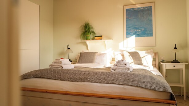 Een opgemaakt bed met handdoeken, een bedlamp, nachtkastje, een plant en een schilderij aan de muur.