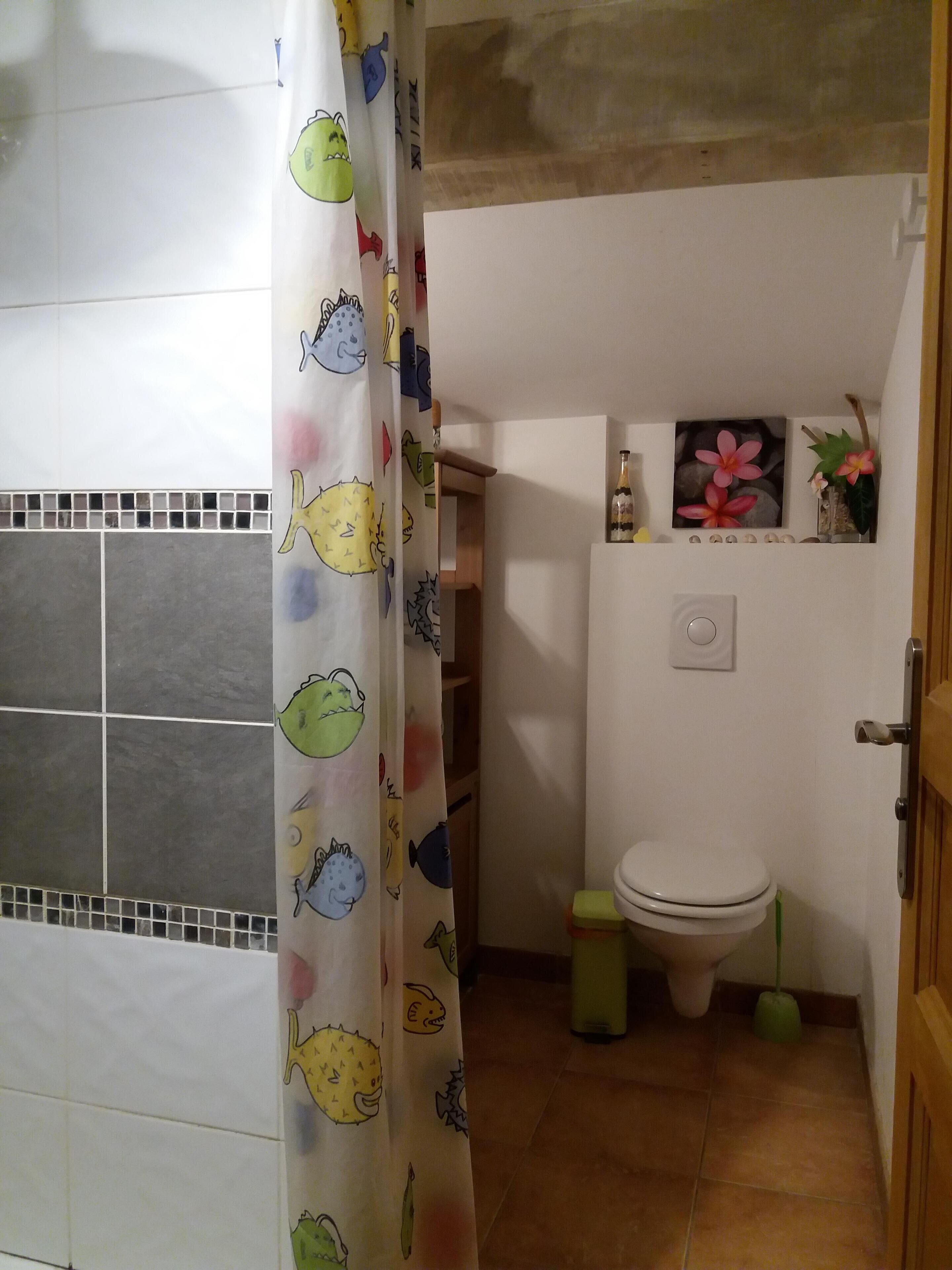 A bathroom with a shower curtain featuring cartoon fish, a toilet, a small shelf with decorative items, and a tiled floor.