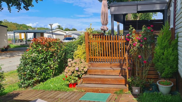 Ein hölzernes Tor führt zu einem Haus mit einer Veranda, umgeben von verschiedenen Kübelpflanzen und Blumen.