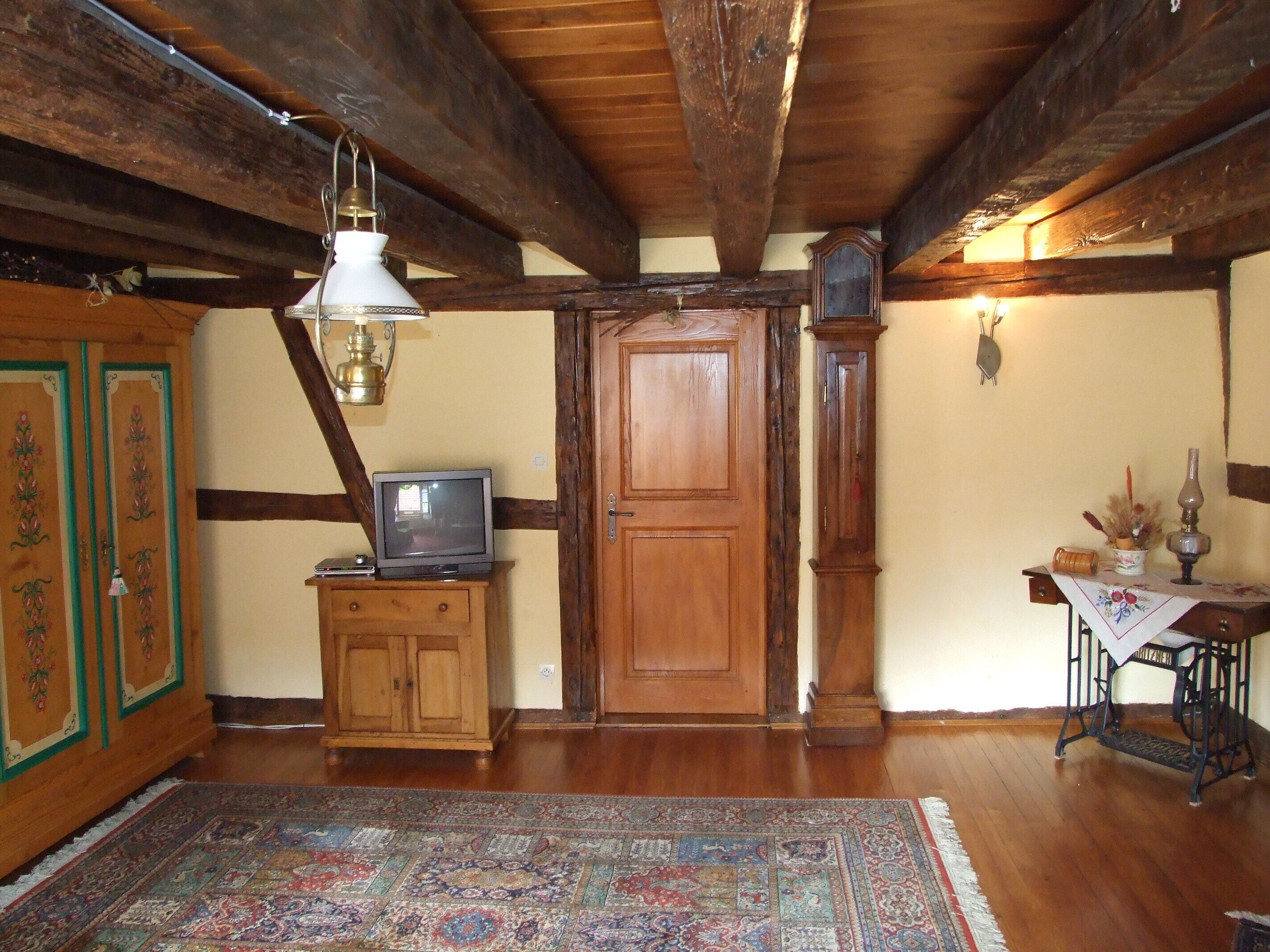 A traditional room with wooden beams, a wooden door, a television on a cabinet, a sewing machine, and a patterned rug.