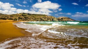 A beach with waves, a sandy shore, and a hill with buildings in the background. - CALM - ZENITUDE - ONE OF THE MOST BEAUTIFUL HOUSES OF GOZO (GHARB)