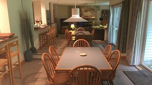 Un coin repas avec une table rectangulaire et six chaises en bois, un luminaire suspendu au-dessus, et une cuisine en arrière-plan.