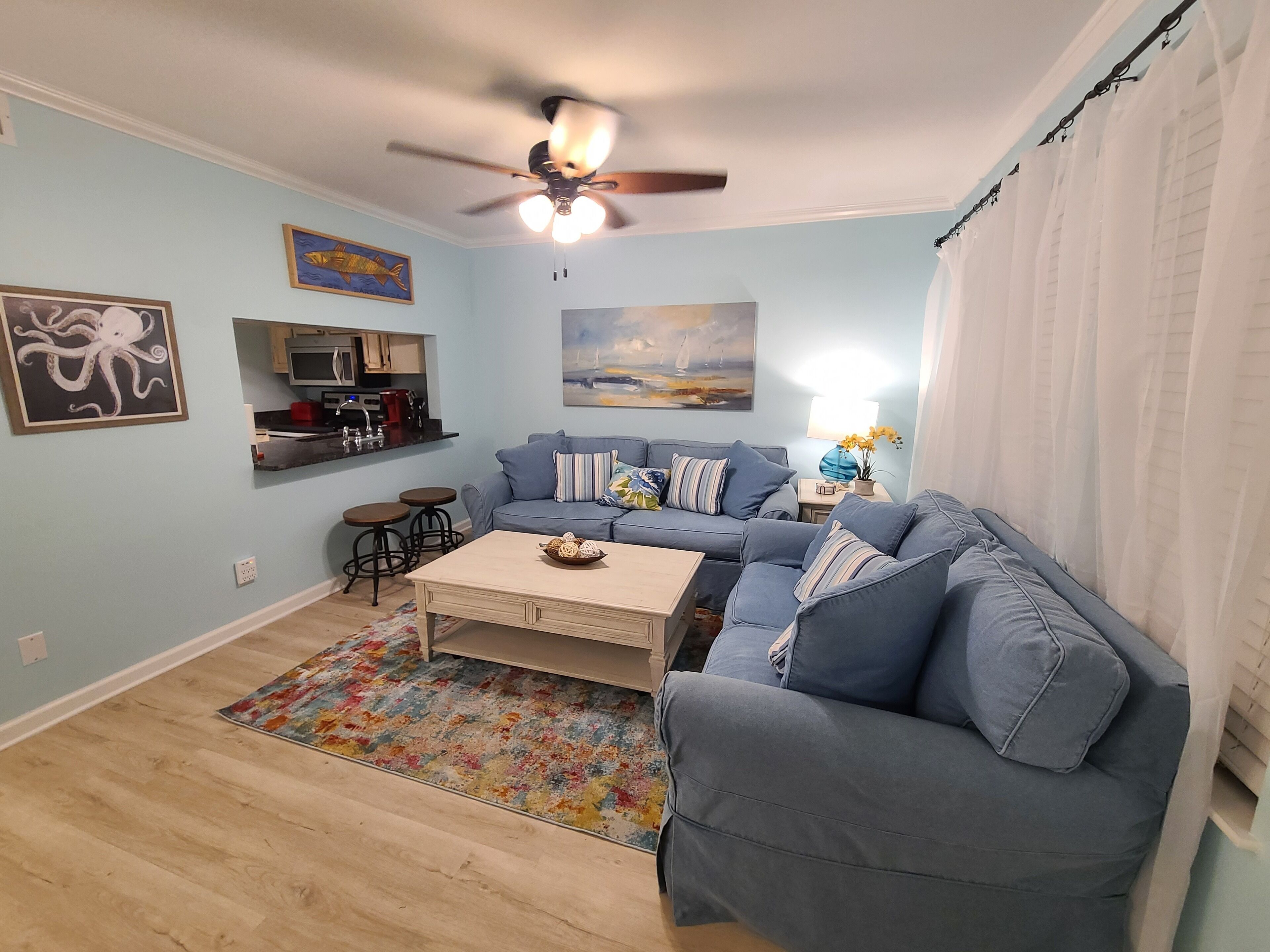A living room with a blue sofa, a coffee table, a ceiling fan, and a kitchen area in the background.