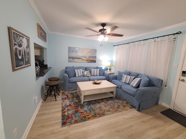 A living room with a blue sofa set, a coffee table, a ceiling fan, and a colorful rug. - Beautiful Condo Steps to the Gulf!  (Gulf Shores)