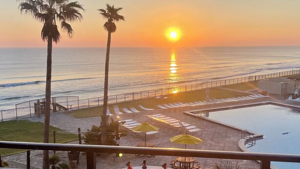 Un balcón con vista a la puesta de sol sobre el océano, una piscina y sillas de descanso.
