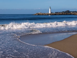 Une plage où les vagues se brisent près du rivage, un phare en arrière-plan et une digue rocheuse.