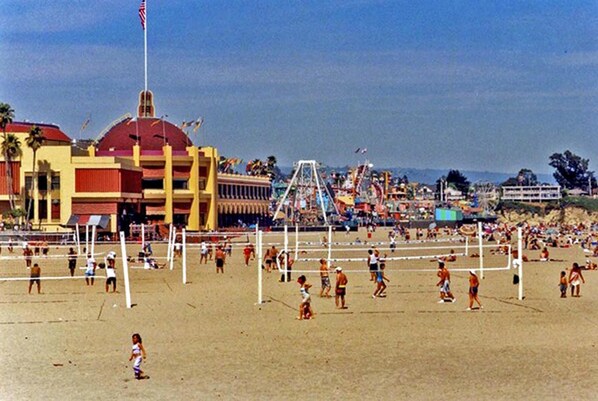 Uma quadra de voleibol de praia com jogadores, um edifício grande com telhado vermelho e uma roda gigante ao fundo.
