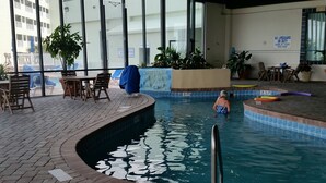 Una piscina interior con borde de azulejos, una estación de salvavidas y una zona junto a la piscina con sillas y mesas.
