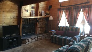 A cozy cabin interior with a stone fireplace, wooden walls, a television, and plaid-patterned furniture. - Sugarloaf Cabin sweet cabin (Big Bear)