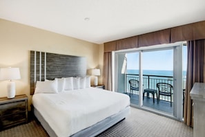 A hotel room with a large bed, a nightstand with a lamp, a view of the ocean through a sliding glass door, and a balcony with chairs. - 4BR/3BA, Sleeps 16, Large Oceanfront, Balcony (Myrtle Beach)