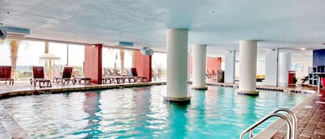 Una piscina interior con agua cristalina, rodeada de sillones de descanso y una zona techada.