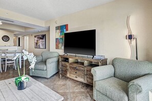 A living room with a flat-screen TV, two grey sofas, a wooden coffee table, and a dining area with a white table and chairs. - 4BR/3BA, Sleeps 16, Large Oceanfront, Balcony (Myrtle Beach)