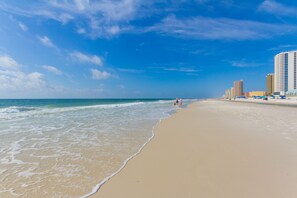 A wide sandy beach with gentle waves, a clear blue sky, and high-rise buildings in the background. - Fenced yard, loveDogs,  Wonderful  Rustic cottage,  Perfect Location!! (Gulf Shores)