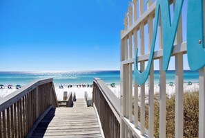 A wooden boardwalk leading to a beach with a clear view of the ocean and a large blue 'M' structure.