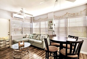 A living room with a sofa, a dining table, and chairs. There is a ceiling fan, a window with blinds, and a decorative starfish garland on the wall. - 50 Steps to Beach Villa Hot Tub 1bd modern cottage (Bradenton Beach)