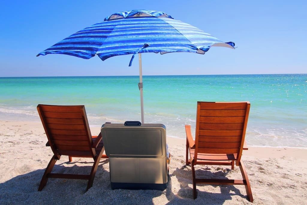 Two wooden lounge chairs and a suitcase on a sandy beach with a blue and white striped umbrella providing shade.