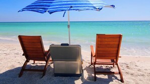 Deux fauteuils de plage en bois et une valise sur une plage de sable, sous un parasol bleu et blanc à rayures qui offre de l’ombre.