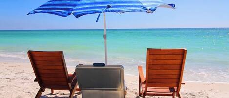 Dos sillones de madera y una maleta sobre una playa arenosa, con una sombrilla azul y blanca que da sombra.