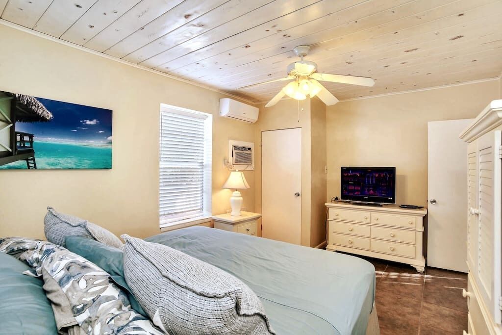 Une chambre à coucher comprenant un lit, un ventilateur de plafond, un téléviseur, une commode et un tableau représentant une scène de plage.