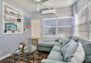 A living room with a glass coffee table, a gray sofa, and a ceiling fan. There are shelves with decorative items and a sign that reads "Our Beach House".