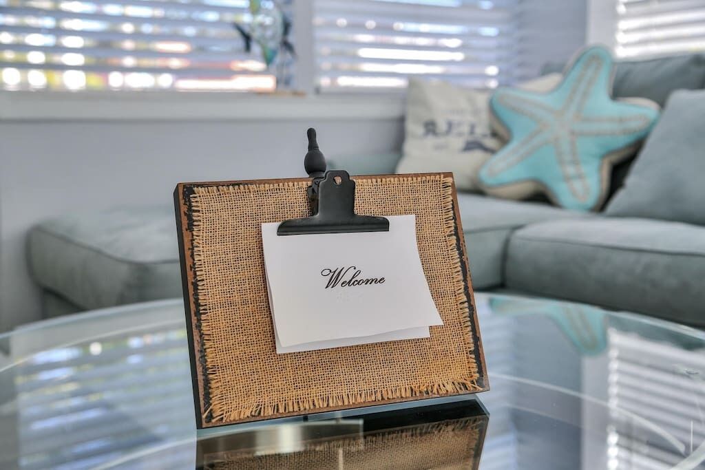 A framed welcome sign on a glass table in a living room with a starfish pillow and a grey sofa.