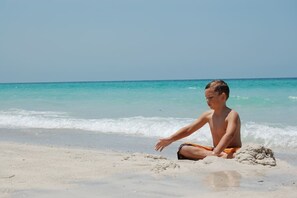 Eine Person sitzt am sandigen Strand, im Hintergrund das Meer.