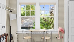 Quarto com uma mesa redonda e duas cadeiras, uma porta branca e uma janela com vista para árvores.