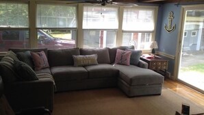 A living room with a sectional sofa, a ceiling fan, large windows, and a view of a red car outside.