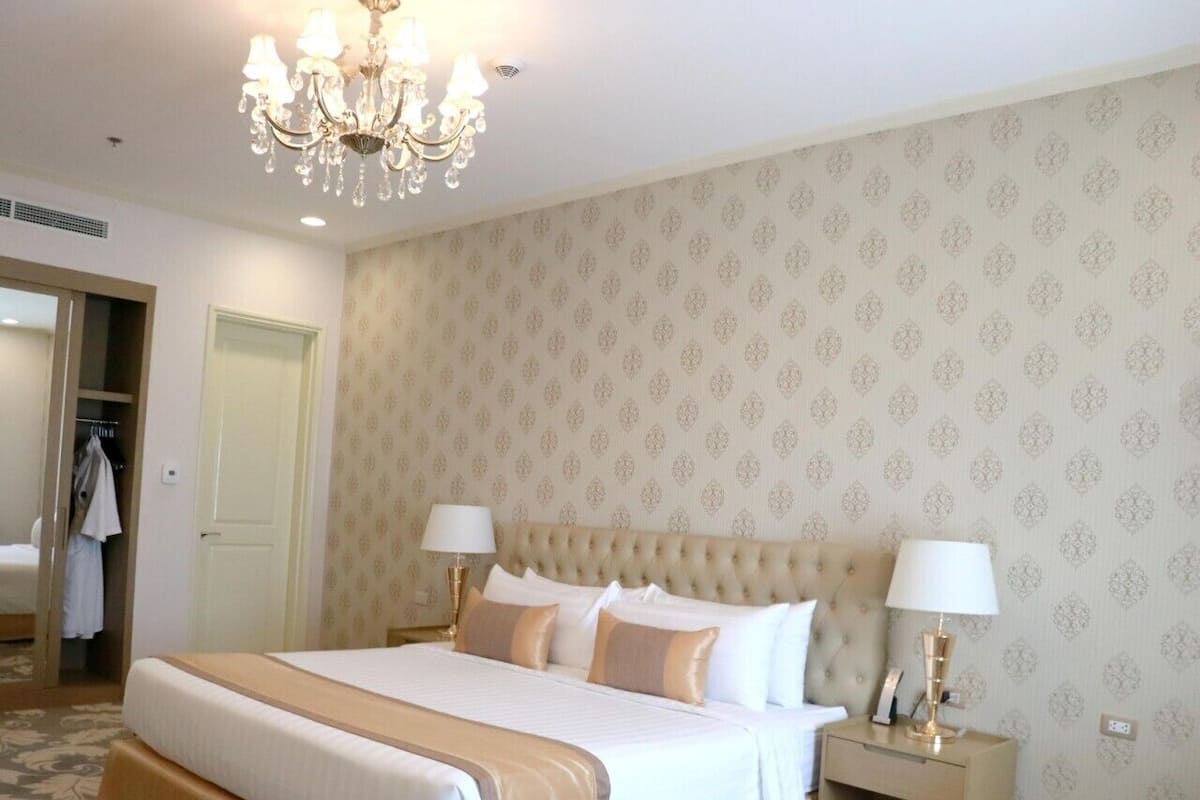Un grand lit avec une tête de lit beige, deux lampes de chevet et un lustre.