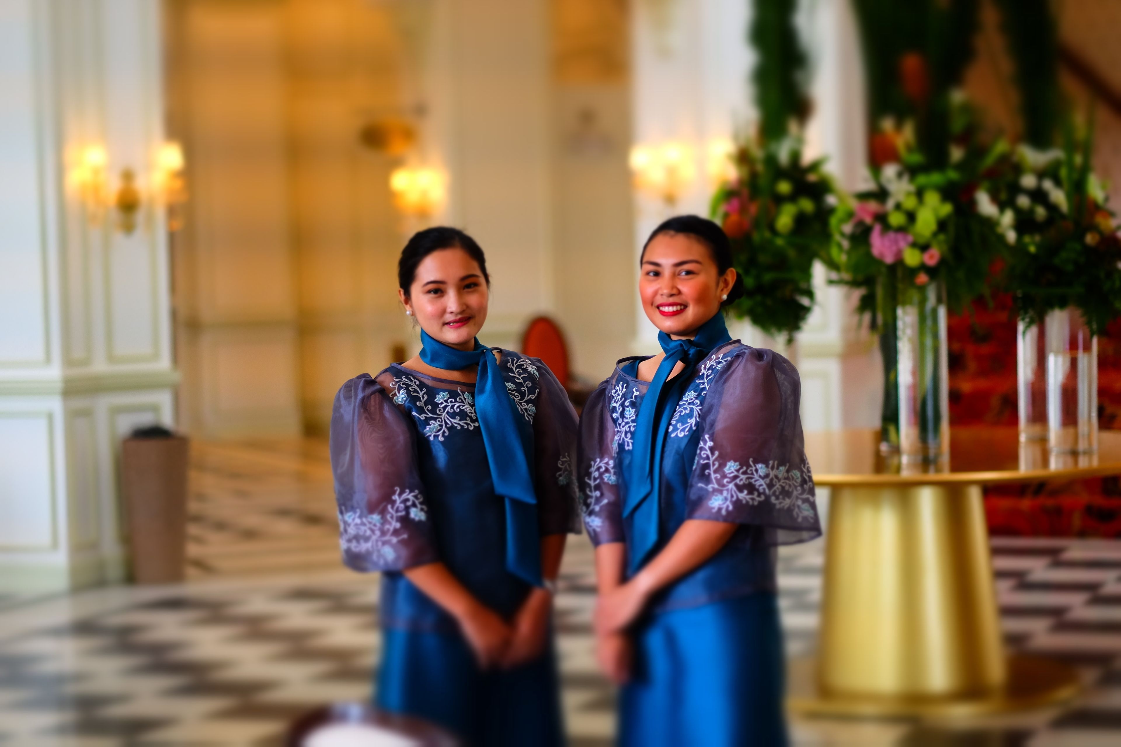 Twee personen in blauwe jurken met gedetailleerd borduurwerk, staan in een goed verlichte kamer met een geruite vloer en een gouden decoratiestuk op tafel.