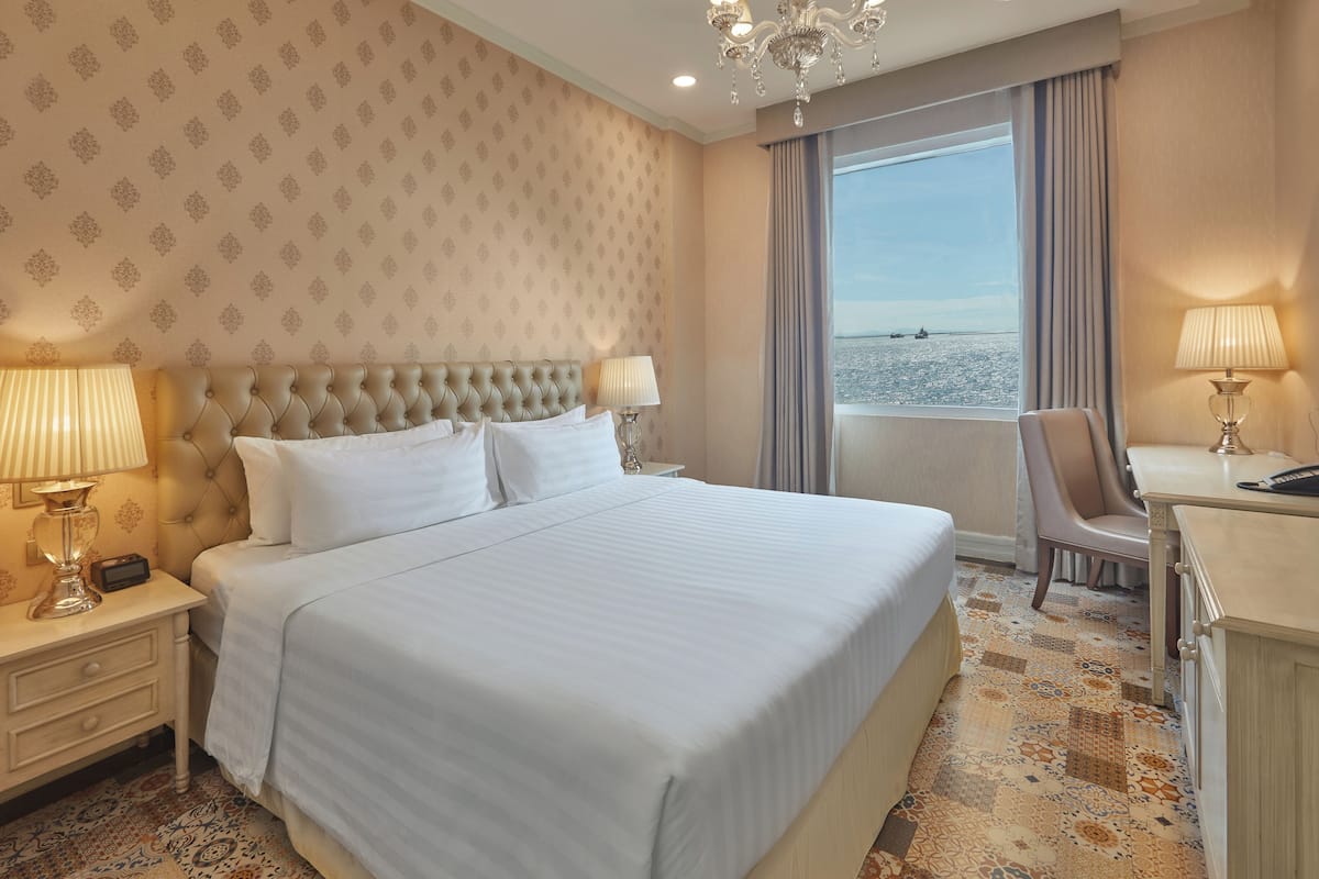 Une chambre d’hôtel avec un grand lit, deux tables de chevet avec des lampes, un bureau avec une chaise et une grande fenêtre donnant sur la mer.