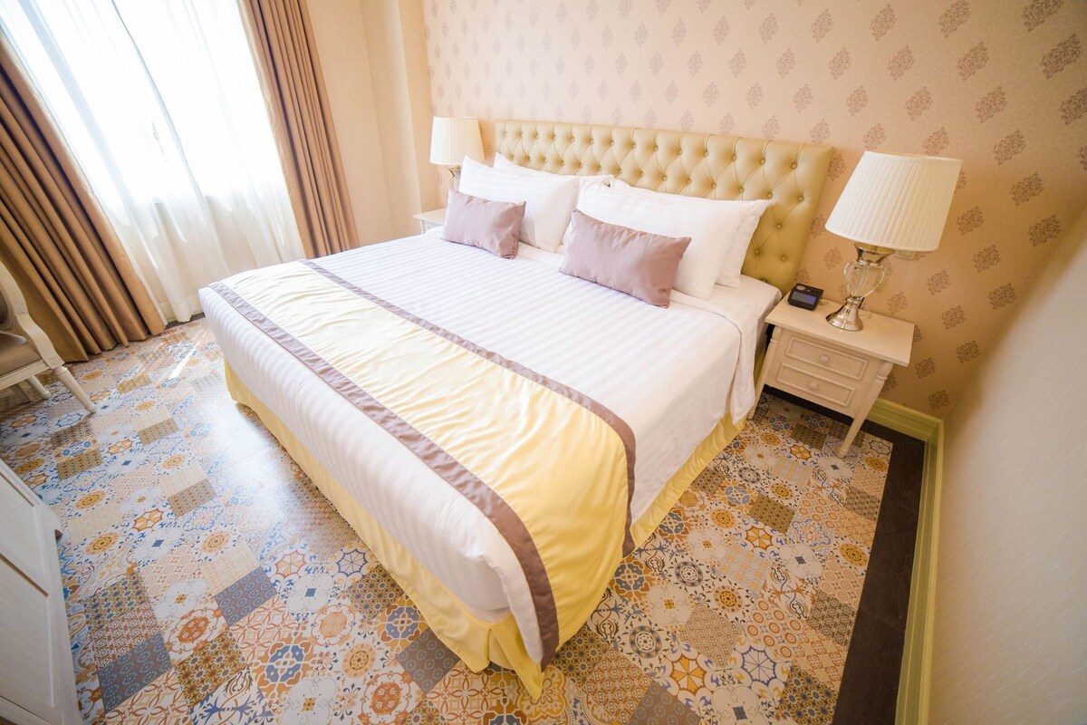 A hotel room with a large bed, a nightstand, a lamp, and patterned floor tiles.