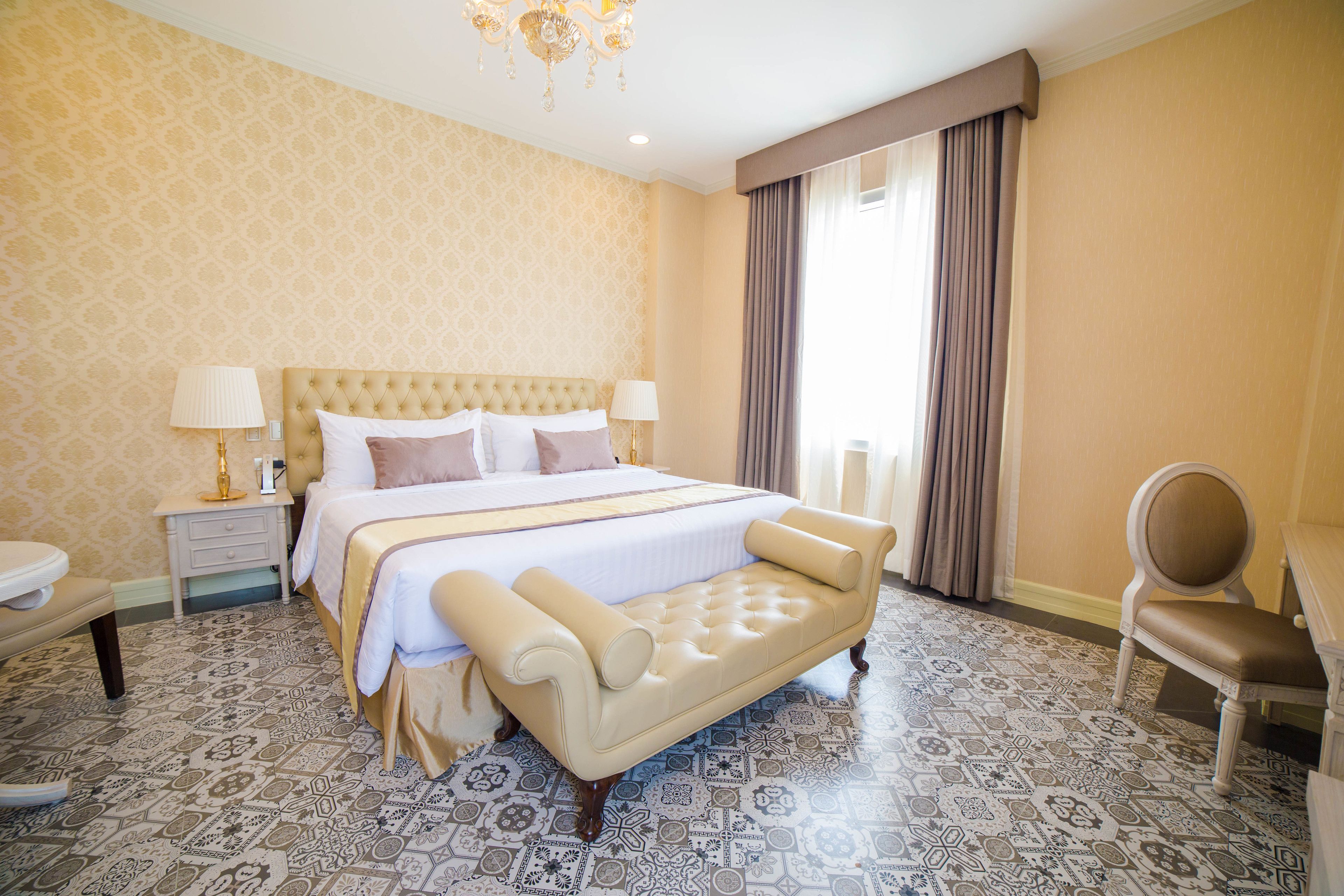 A hotel room with a large bed, a chaise lounge, a chair, a nightstand, and patterned floor tiles.