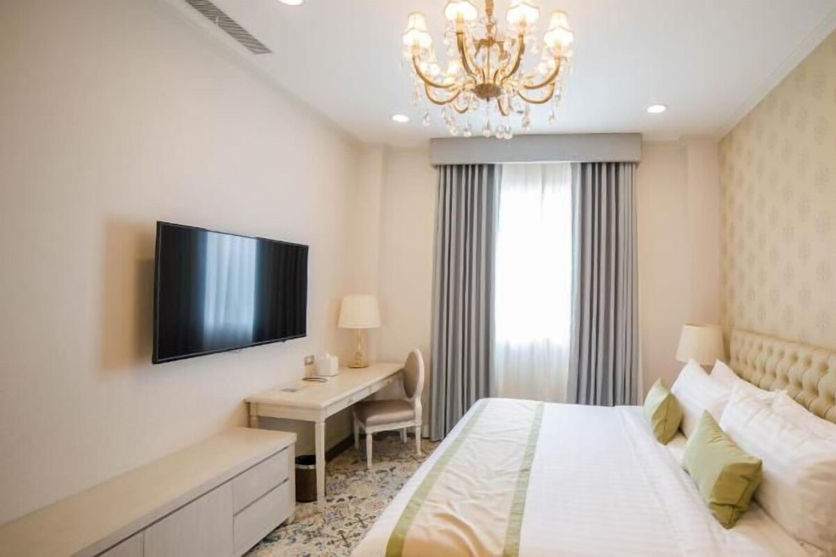 A hotel room with a large bed, a desk, a chair, a television, and a chandelier.